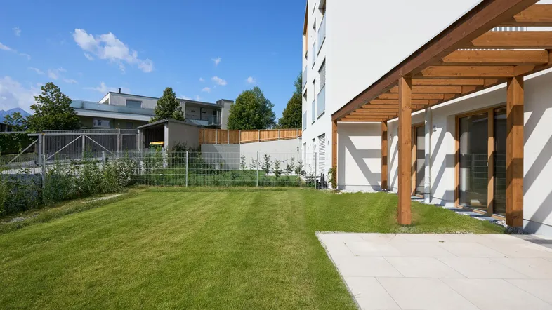 Gepflegter Gartenbereich mit Rasenfläche, heller Terrasse und Holzpergola am modernen Gebäude.