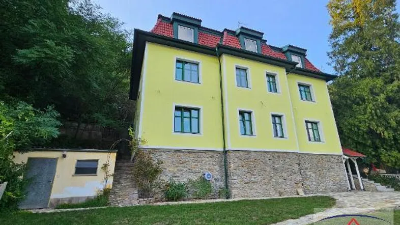 Gelbes Mehrfamilienhaus mit rotem Mansarddach und Steinsockel, umgeben von viel Grün.