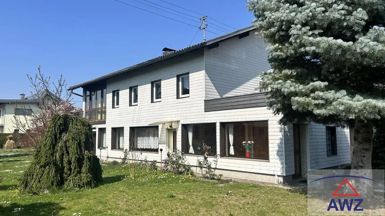 Weißes Einfamilienhaus mit großem Garten und Nadelbaum im Vorgarten unter blauem Himmel.