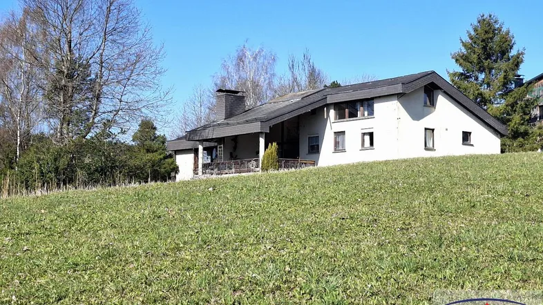 Freistehendes Haus auf einem grünen Hügel mit Bäumen im Hintergrund unter blauem Himmel.