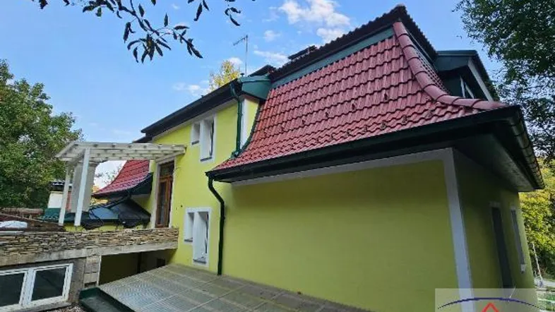 Seitenansicht eines Hauses mit gelber Fassade, rotem Ziegeldach und einer weißen Pergola.
