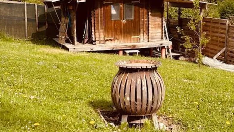Kleines Holzhaus im Grünen mit Feuerstelle und umgebendem Gartenbereich unter blauem Himmel.