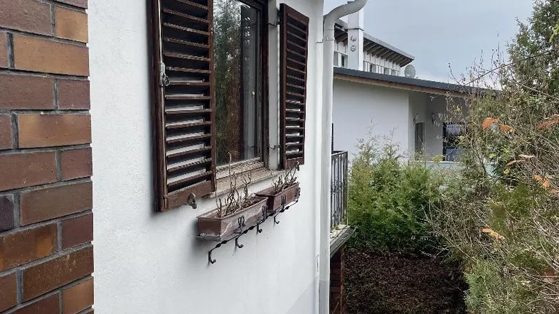Seitenansicht des Hauses mit Fensterläden, Ziegelmauerwerk und einem abfallenden Gartenweg.