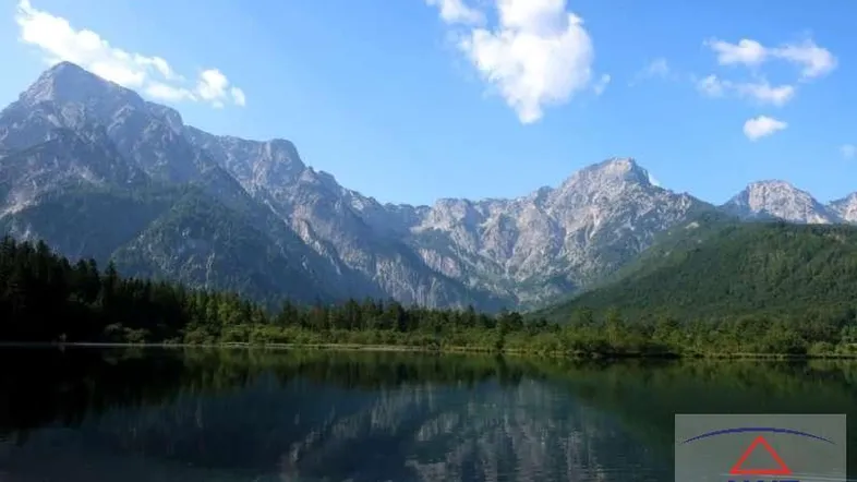 Malerischer Bergsee mit klarer Wasseroberfläche, umgeben von majestätischen Bergen und grünen Wäldern unter blauem Himmel.