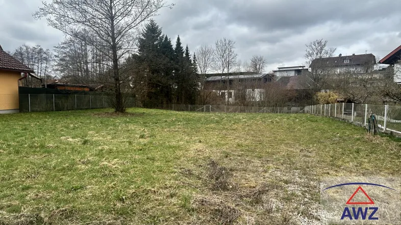 Leeres Grundstück mit Grasfläche, einigen Bäumen und Häusern im Hintergrund unter bewölktem Himmel.