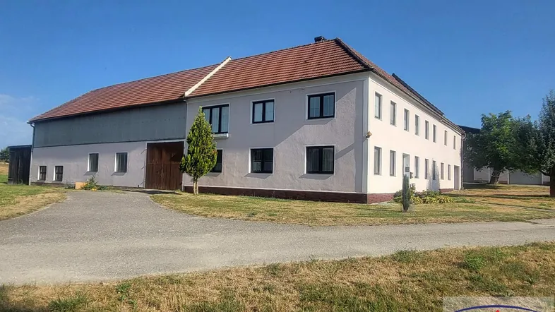 Großes Gebäude mit heller Fassade, rotem Ziegeldach und angrenzendem Scheunenteil in ländlicher Umgebung.