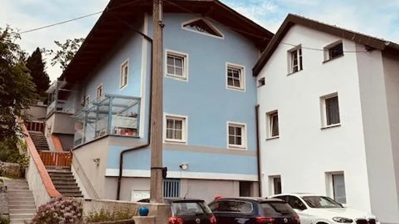 Zwei Wohnhäuser mit hellblauer und weißer Fassade und Parkplätzen davor unter bewölktem Himmel.