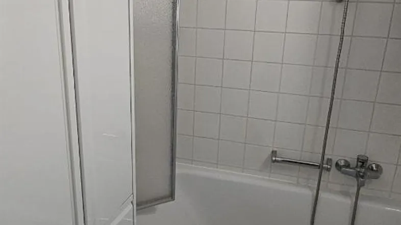 Badezimmer mit Badewanne und Duschvorrichtung, weißen Fliesen und praktischem Stauraum.