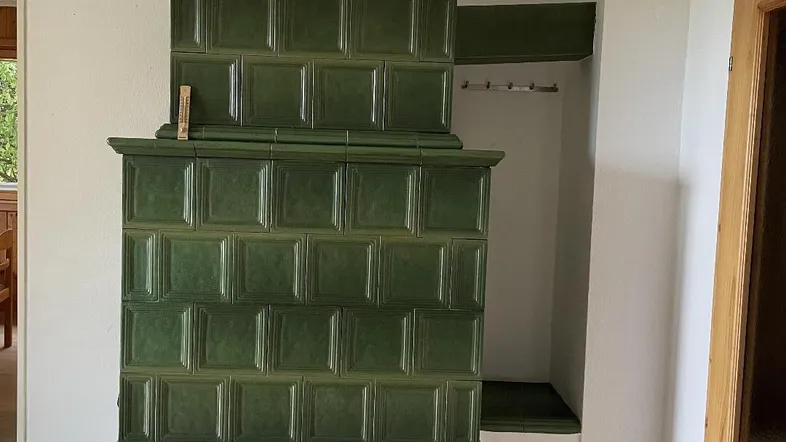 Traditioneller grüner Kachelofen in einem hellen Wohnbereich mit Holzboden und Zugang zum Außenbereich.