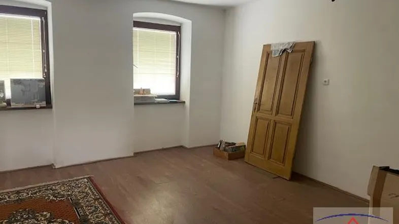Leerer Raum mit Holzboden, zwei Fenstern mit Jalousien und einer Holztür, die an der Wand lehnt.