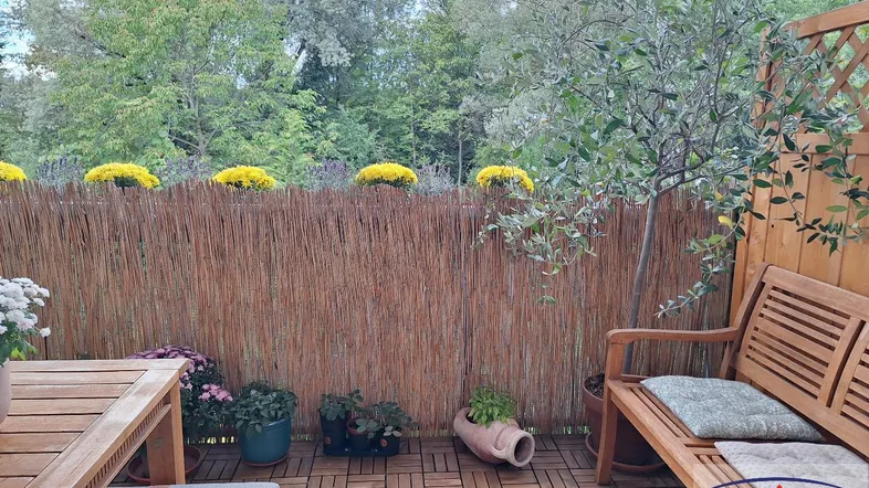 Gemütliche Terrasse mit Holzboden, Sitzgelegenheiten und üppiger grüner Bepflanzung im Hintergrund.