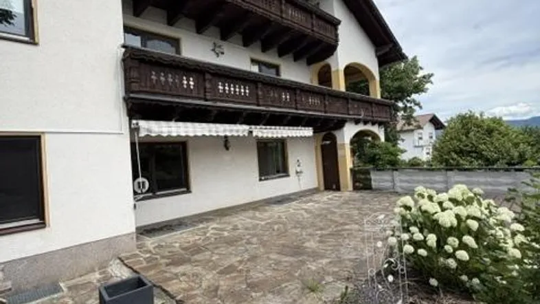 Traditionelles Haus mit Holzbalkonen und gepflastertem Vorplatz, umgeben von Grün.
