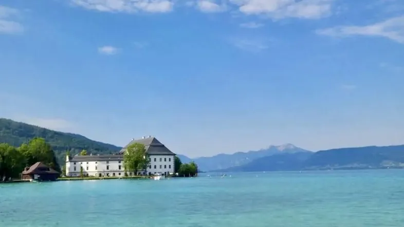 Historisches Gebäude am Ufer eines klaren Sees mit Bergen im Hintergrund unter strahlend blauem Himmel.