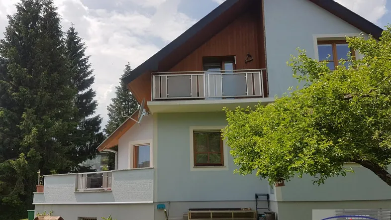Ansicht des Hauses mit Balkon, kleiner Terrasse und umgebender Grünfläche unter bewölktem Himmel.
