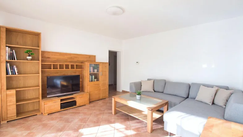 Helles Wohnzimmer mit beigem Fliesenboden, grauem Ecksofa und Holzmöbeln.