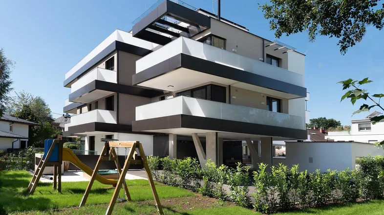 Modernes Mehrfamilienhaus mit gestuften Balkonen, Spielplatz und gepflegter Grünfläche unter blauem Himmel.