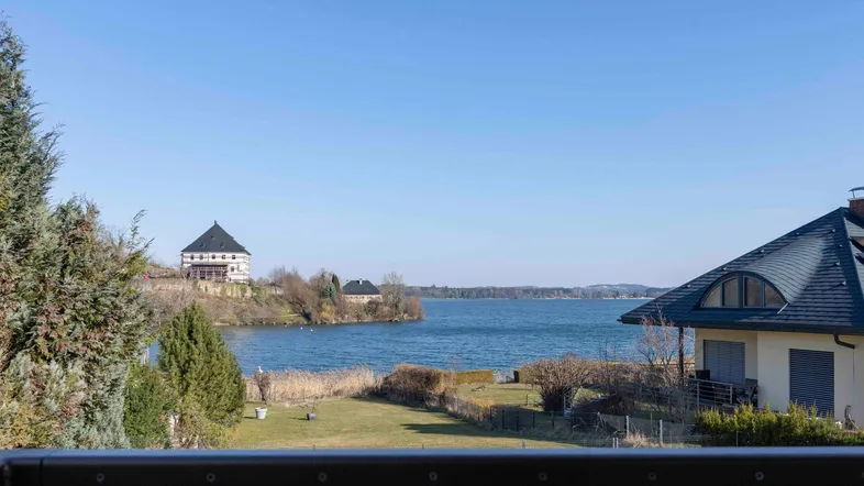 Blick vom Balkon auf einen malerischen See mit umliegender Natur und Gebäuden am gegenüberliegenden Ufer.