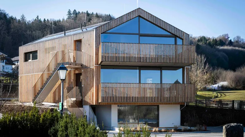Modernes Mehrfamilienhaus mit Holzfassade, großen Glasflächen und Balkonen in ländlicher Umgebung.