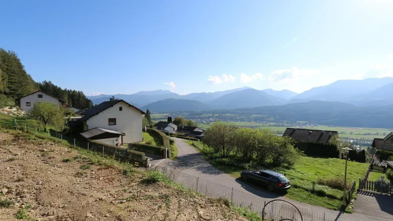 Panoramablick auf Häuser in einem Tal mit Bergen im Hintergrund unter blauem Himmel.