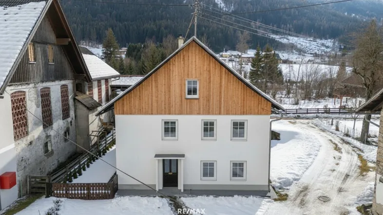 Traditionelles Haus mit weißer Fassade und Holzgiebel in einer verschneiten Berglandschaft, umgeben von Natur.