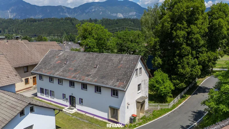 Luftaufnahme eines Wohnhauses mit Satteldach und umliegender grüner Landschaft sowie Bergen im Hintergrund.
