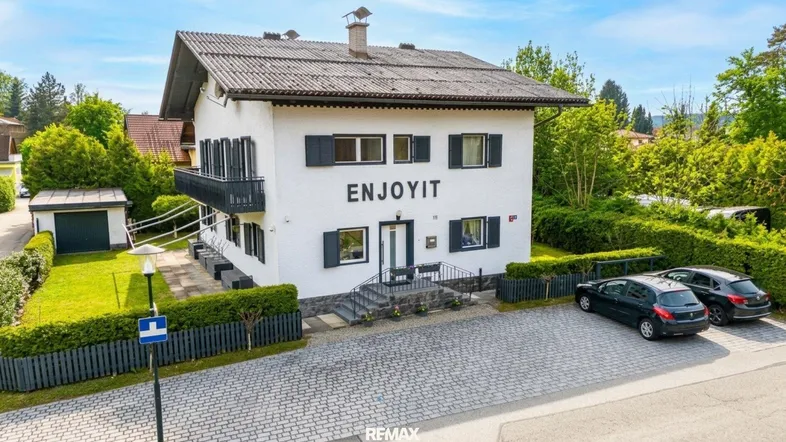 Weißes Haus mit dunklem Dach, Fensterläden und gepflastertem Vorplatz mit Parkplätzen.