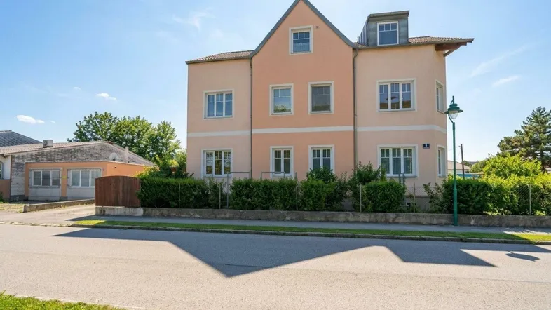 Mehrstöckiges Gebäude mit hellrosa Fassade und gepflegtem Vorgarten an einer ruhigen Straße.