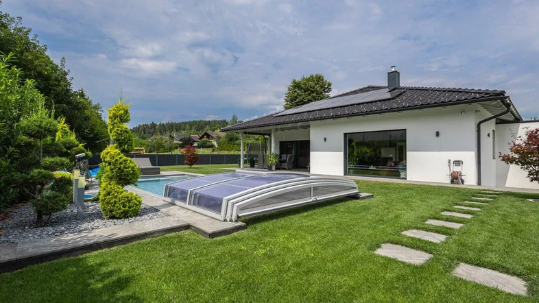 Großzügiger Gartenbereich mit Rasenfläche, Pool und moderner Hausfassade unter blauem Himmel.