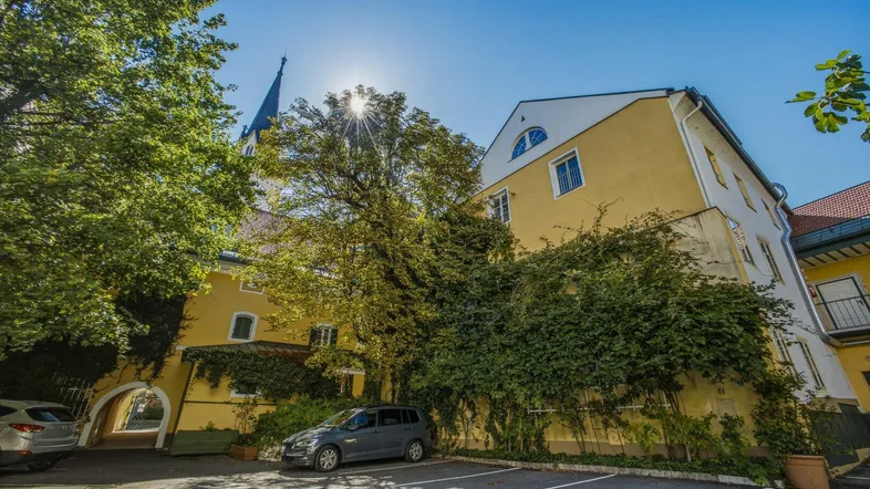 Gelbes historisches Gebäude mit Parkplatz und üppiger Begrünung unter blauem Himmel.