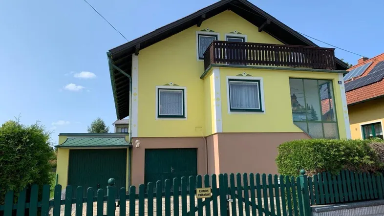 Gelbes Einfamilienhaus mit Balkon, Garage und grüner Umzäunung unter blauem Himmel.