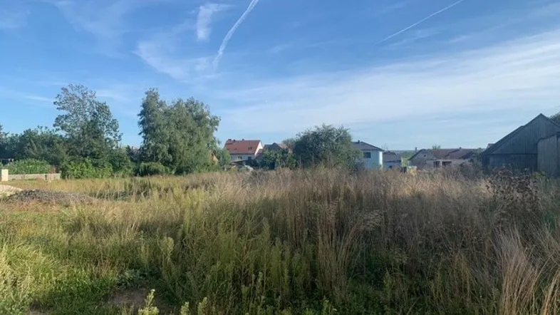 Weitläufiges Grundstück mit hohem Gras und Häusern im Hintergrund unter blauem Himmel.