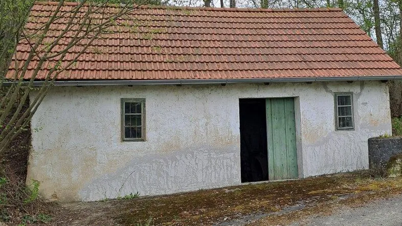 Altes, einfaches Gebäude mit verputzten Wänden, einem Ziegeldach und grünen Holztüren, umgeben von Bäumen.