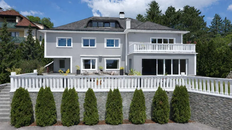 Moderne Villa mit grauer Fassade, weißem Balkon und gepflegter Gartenanlage.