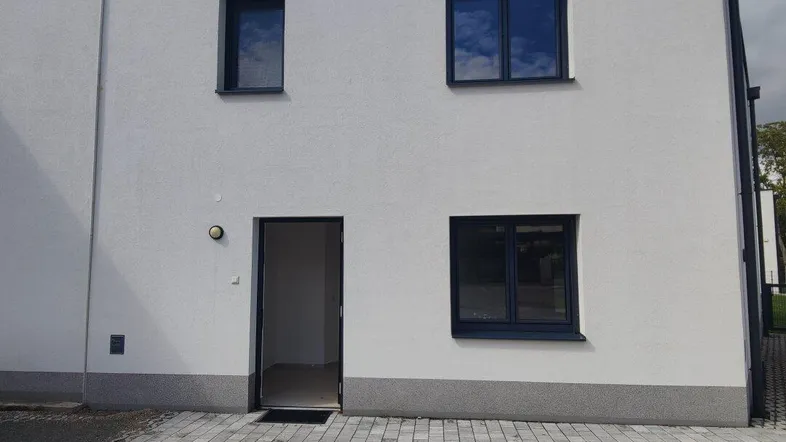 Moderne Hausfassade eines Neubaus mit gepflastertem Eingangsbereich und dunklen Fenstern.