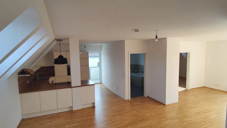 Heller offener Wohnbereich mit Holzboden, Dachschrägen und integrierter Küchenzeile in einer Dachgeschosswohnung.