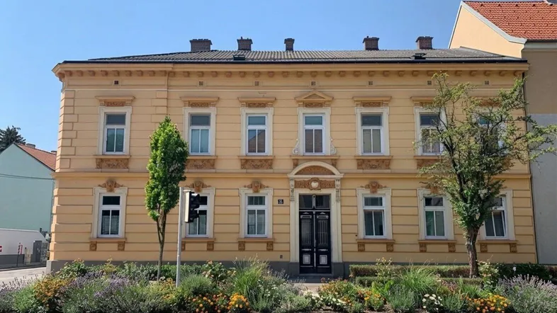 Klassisches gelbes Gebäude mit reich verzierter Fassade und einem gepflegten Vorgarten unter blauem Himmel.