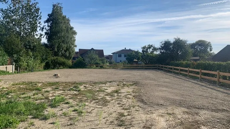Leeres Grundstück mit Holzumzäunung und Blick auf umliegende Wohnhäuser unter blauem Himmel.