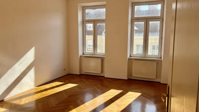 Helles Zimmer mit Fischgrätparkett und zwei Fenstern, durch die Sonnenlicht einfällt.