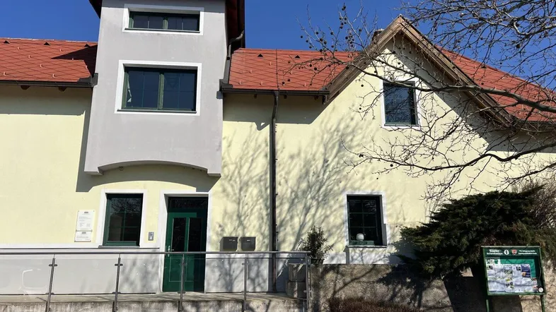 Gelbe Fassade eines Gebäudes mit grauem Anbau und rotem Ziegeldach unter blauem Himmel.