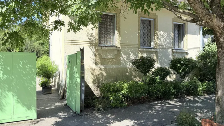 Vorderansicht des Hauses mit grünen Fensterläden und einem gepflegten Vorgarten unter schattenspendenden Bäumen.