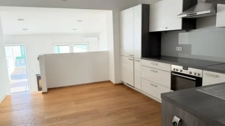 Moderne Küche mit hellen Schränken, dunkler Arbeitsplatte und Holzboden, offen zu einem weiteren Wohnbereich.