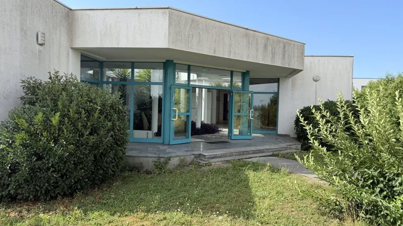 Eingangsbereich eines Gebäudes mit großen Glasfronten und umliegender Grünfläche unter blauem Himmel.