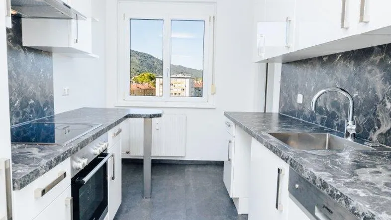 Moderne weiße Küche mit grauen Arbeitsplatten, integrierten Geräten und Bergblick durch das Fenster.