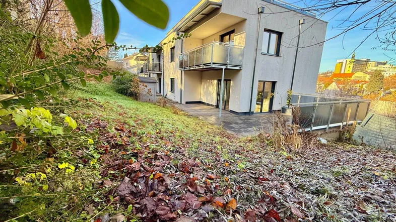 Modernes Mehrfamilienhaus mit Balkonen und Terrasse, eingebettet in eine grüne Hanglage.