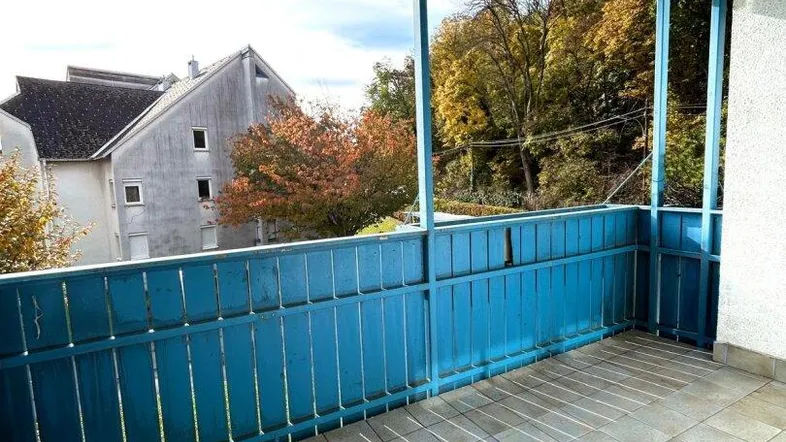Heller Balkon mit blauer Brüstung und Blick auf umliegende Häuser und Bäume.