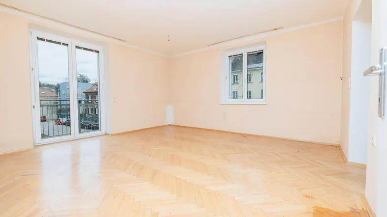 Helles, leeres Wohnzimmer mit Fischgrätparkett und Zugang zu einem Balkon.