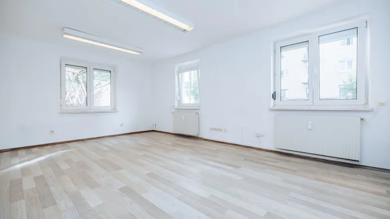 Helles, leeres Zimmer mit weiß gestrichenen Wänden, Laminatboden und mehreren Fenstern.