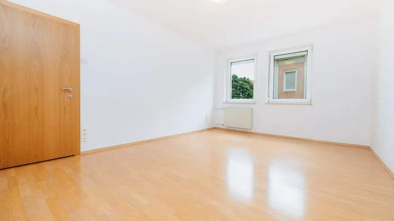 Helles, leeres Zimmer mit Laminatboden und zwei Fenstern, bereit zur individuellen Gestaltung.