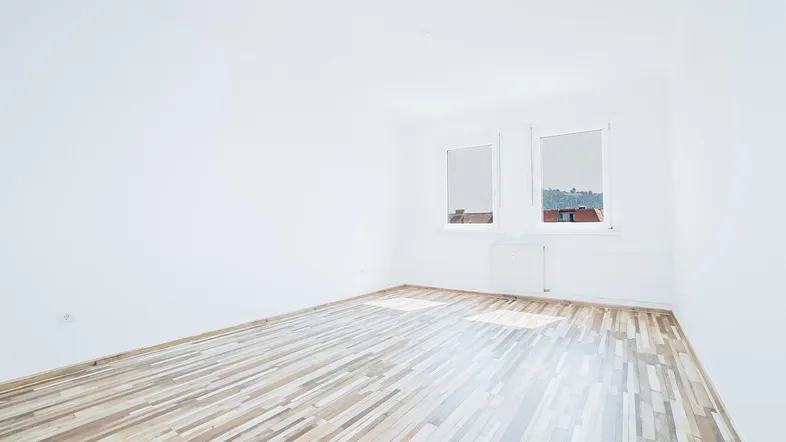 Helles, leeres Zimmer mit Laminatboden und zwei Fenstern, das viel Tageslicht hereinlässt.