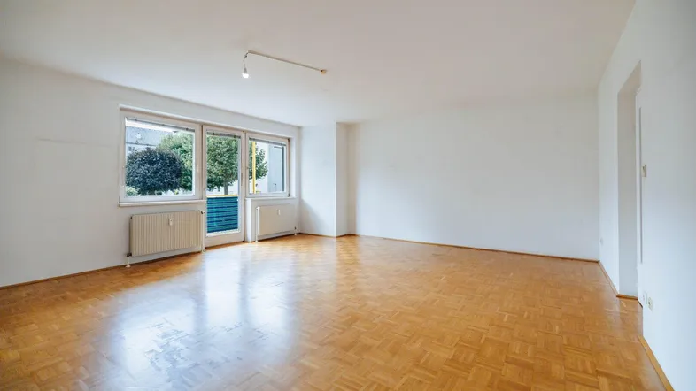 Helles, leeres Wohnzimmer mit Fischgrätparkett und Zugang zu einem Balkon oder einer Terrasse.